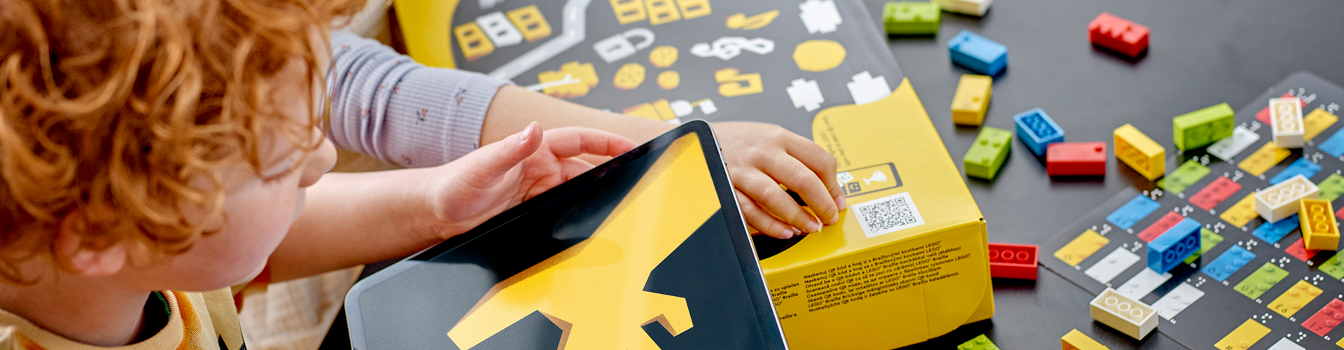 Ein Kind hält einen schwarz-gelben Karton in der Hand, um es herum sind bunte LEGO Steine, die mit Braille Schrift bedruckt sind.